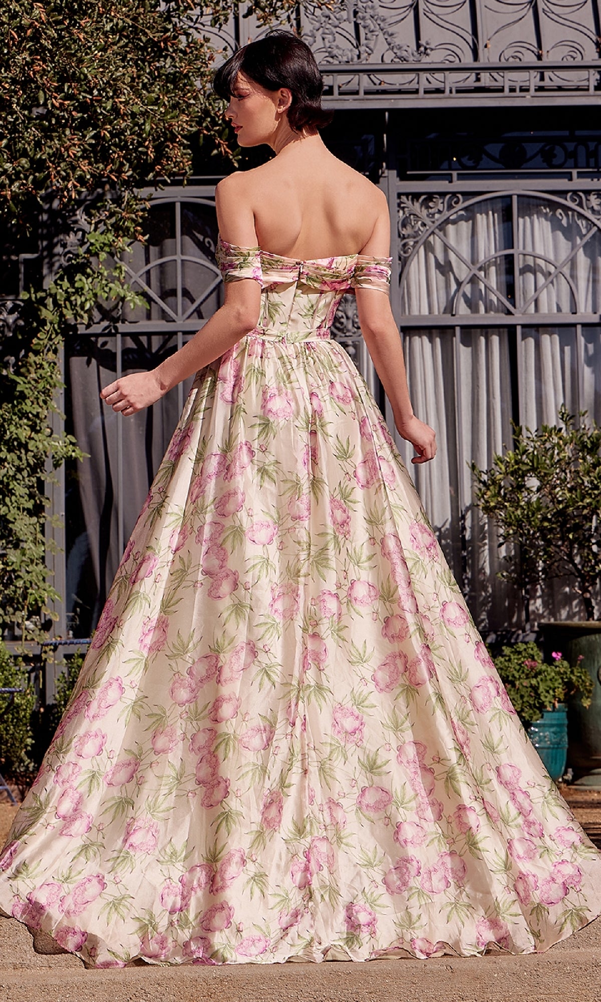 A woman stands outdoors, facing away from the camera, wearing the Andrea & Leo A1466 long floral-print prom dress featuring an off-the-shoulder design and corset-style bodice.